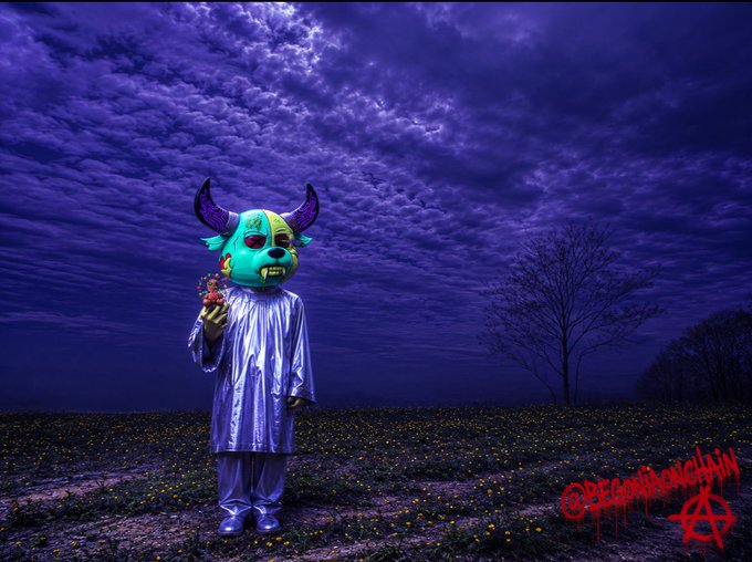A surreal dark fantasy image of a horned, fanged figure in a shiny purple robe holding flowers in a field under a dramatic purple cloudy sky, tagged '@BeGotBlacklight'.