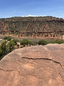 A wide desert landscape featuring layered red rock formations, sparse green vegetation, and a prominent flat sandstone surface in the foreground under a partly cloudy sky.