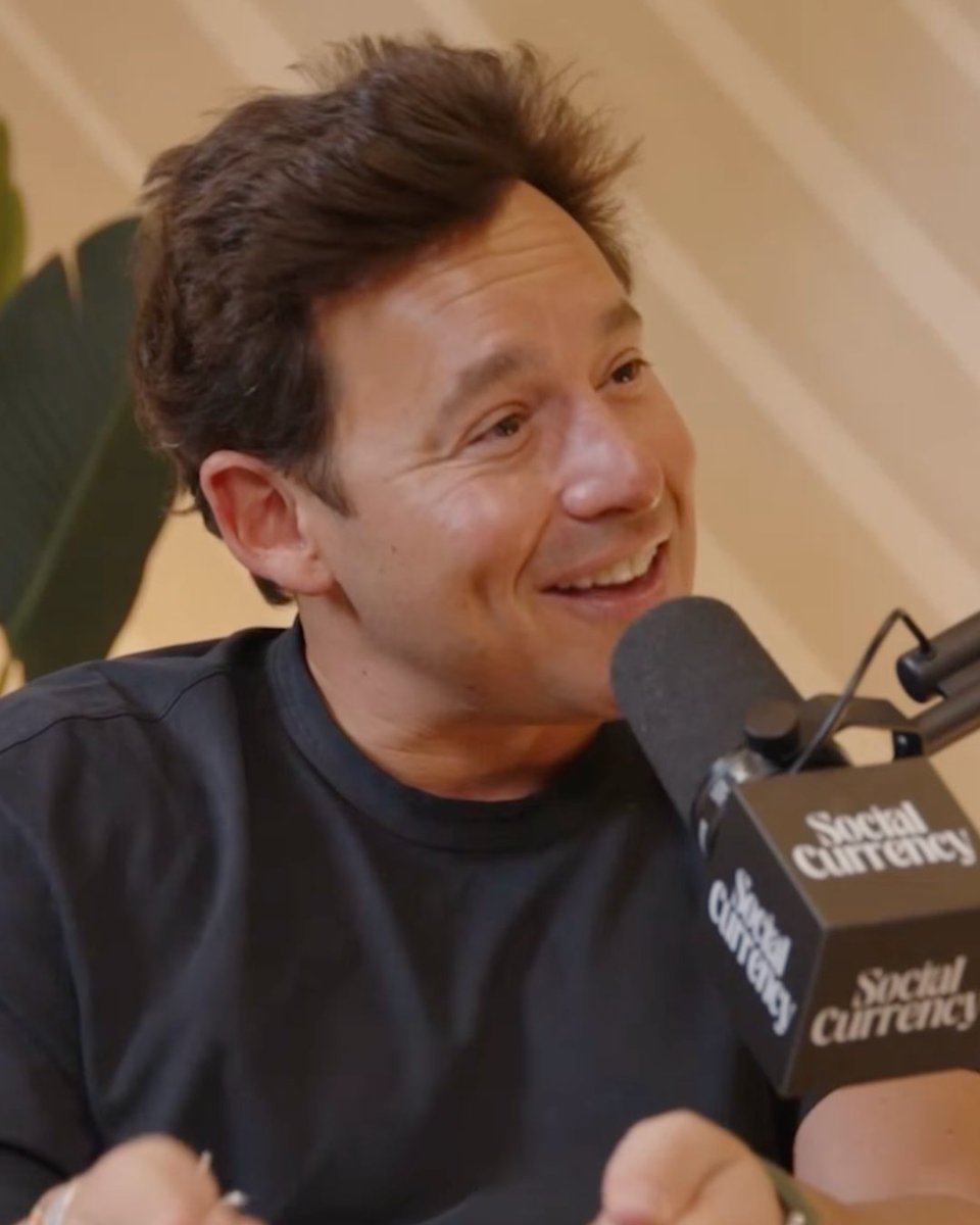 The image features a man wearing a black shirt, sitting down with his hands behind his back while smiling at the camera. The background includes a wall and a plant, creating an indoor setting for the scene. There are also some text elements in the image that seem to be related to social media or online content, such as "Social Currency" and "x402".