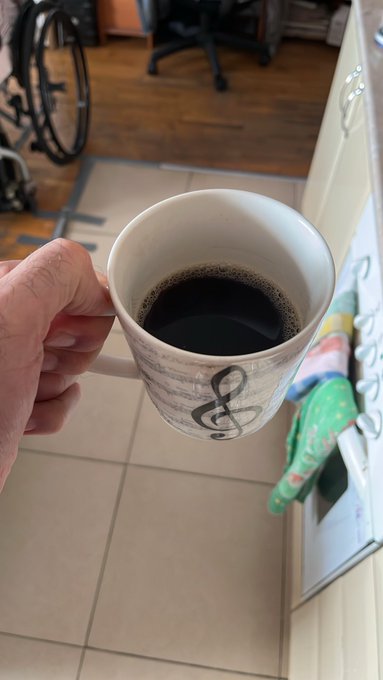 A first-person view of a hand holding a white mug filled with black coffee, decorated with musical notes and a treble clef.