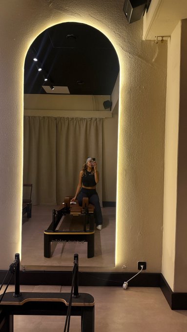 A person takes a mirror selfie in a dimly lit room featuring a large arched mirror with warm backlighting, with Pilates reformer equipment visible in the foreground and reflection.