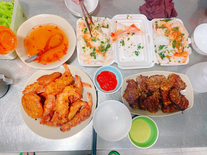 An overhead view of a meal spread on a metal table includes fried shrimp, fried meat, rice with herbs, chili sauce, and a bowl of orange dipping sauce.