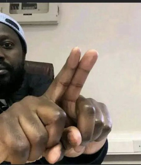 A close-up video still of a man with a beard and a gray cap holding up two fingers, with the fingers appearing warped and distorted, possibly due to a visual filter.