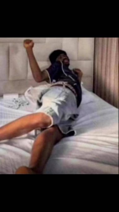 A person wearing a black beanie and dark jacket lying on a white bed with one arm raised in the air.