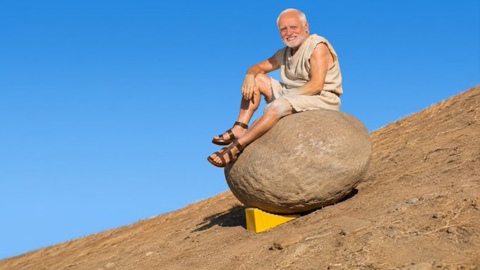 An older man with a white beard, wearing a beige vest and shorts, sits smiling on a large round boulder perched on a sandy hill under a blue sky.