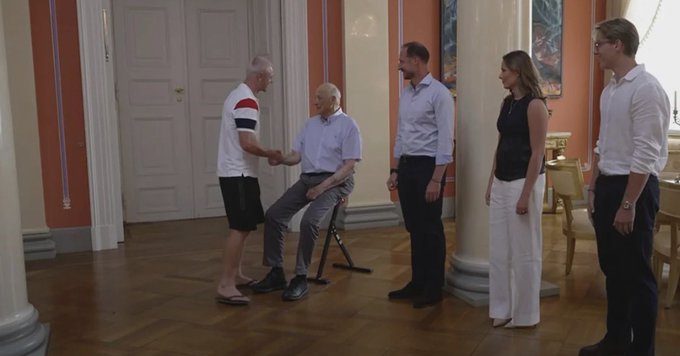 A man wearing flip-flops, a white shirt with red and black stripes, and black shorts shakes hands with a seated man in a light blue shirt, while three other people stand nearby in an ornate room with columns, parquet flooring, and pastel-colored walls. The image aligns with the caption, capturing the moment Julian Ryerson arrives at Norway's Royal Palace in Oslo wearing casual flip-flops to meet the seated royal figure.