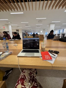 A study space in a library with a laptop, water bottles, and a red 'TAKE A SEAT' card on a wooden desk.