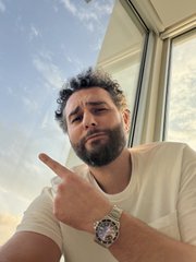A selfie of a bearded man in a white t-shirt pointing at his wristwatch, with a cloudy sky and building visible behind him.