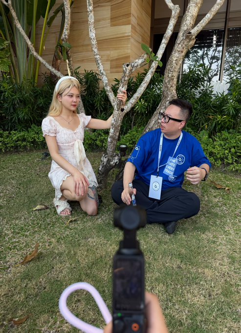 A behind-the-scenes photo showing a blonde woman in a white dress being interviewed by a man in a blue 'Late Show' shirt, filmed by a handheld camera in an outdoor garden setting.