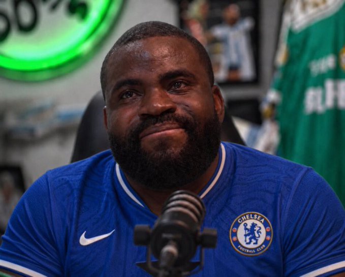 A bearded man wearing a blue Chelsea Football Club jersey sits in front of a microphone in what appears to be a sports podcast setting, with framed photos and a green jersey visible in the background.