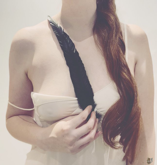 The image shows a woman in a white slip dress, holding a black raven feather against her chest, with her long red hair cascading down. It visually echoes Edgar Allan Poe's "The Raven," specifically the melancholy imagery of Lenore, as the feather serves as a symbol of the poem's haunting, sorrowful tone.