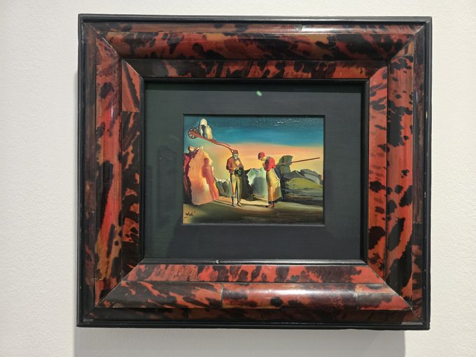 A small framed painting depicting figures and what appears to be a camel against a colorful sky is displayed in a distinctive red and black marbled frame.
