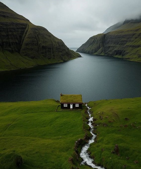 A small grass-roofed house sits on a green hillside with a stream flowing past it, overlooking a fjord surrounded by steep cliffs under an overcast sky.