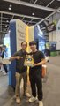 Two young men posing together at what appears to be a gaming or trading card convention, with one holding up a card.