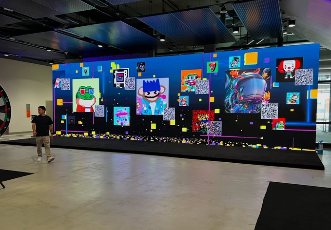 A person walks past a large digital display featuring various pixel art characters, QR codes, and colorful graphics in what appears to be an exhibition space.