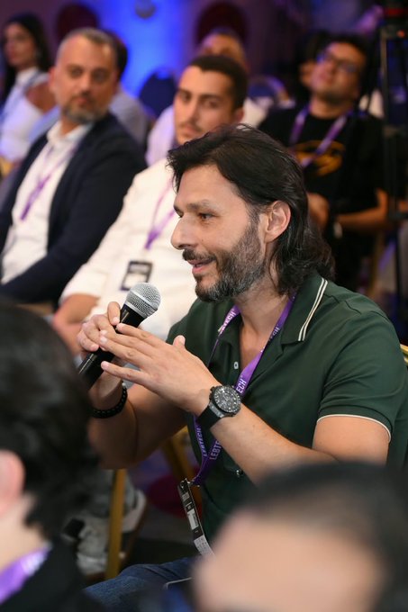 A man with long dark hair and a beard, wearing a green polo shirt and a purple lanyard, speaks into a microphone while seated at an event with other attendees in the background.
