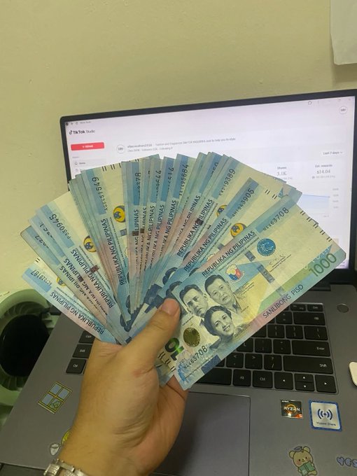 A hand holding a fan of Philippine 100-peso bills in front of an open TikTok Studio dashboard on a laptop.