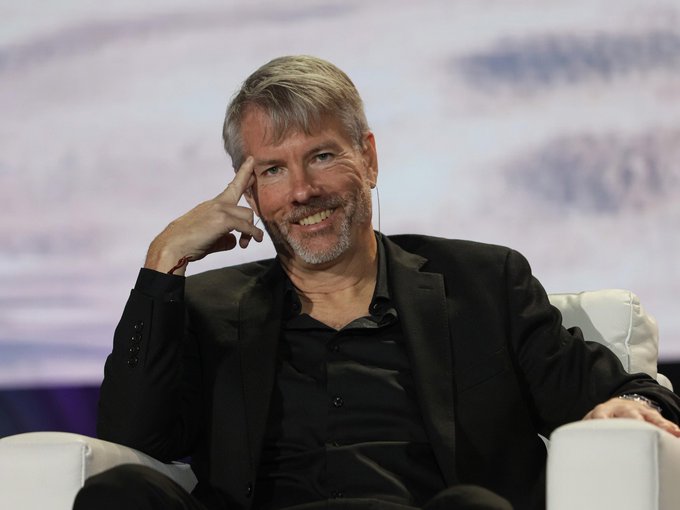 Michael Saylor smiles while seated on a stage, resting his hand against his temple in a thoughtful pose. The image accompanies news of his company, Strategy ($MSTR), increasing its USD reserve by $450 million and now holding 843,775 $BTC alongside $3 billion in cash reserves.