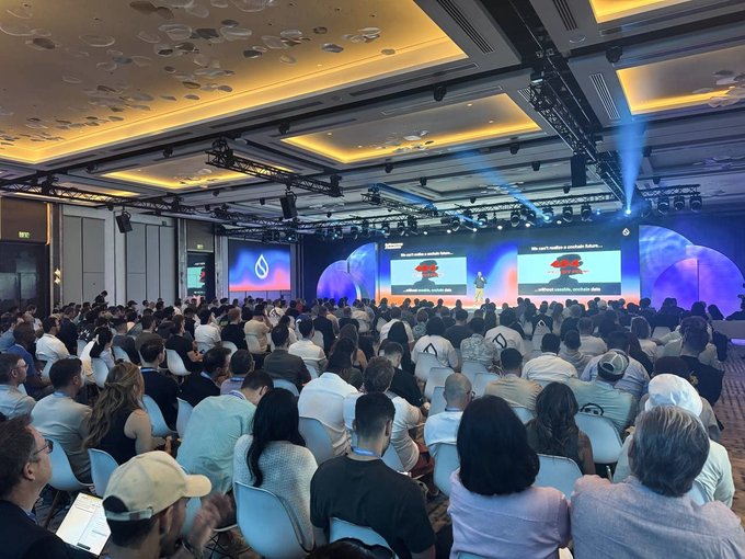A crowded conference room with attendees seated facing a large stage with multiple screens displaying presentation content, lit by stage lights and blue ambient lighting.