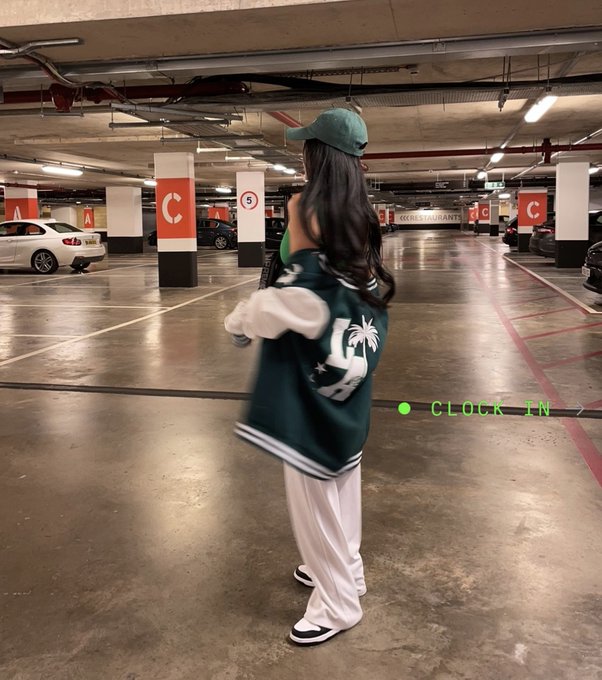 A woman wearing a green cap and varsity jacket stands in a parking garage, with text overlay reading 'CLOCK IN'.