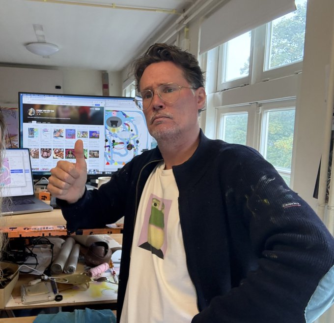 A man wearing glasses, a white t-shirt with a penguin graphic, and a dark jacket gives a thumbs-up in what appears to be an art studio or workspace with computer monitors in the background.