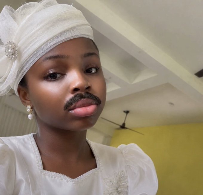 A young person wearing a white traditional headpiece and outfit has drawn on a fake mustache with their tongue sticking out, posing for a selfie-style photo.
