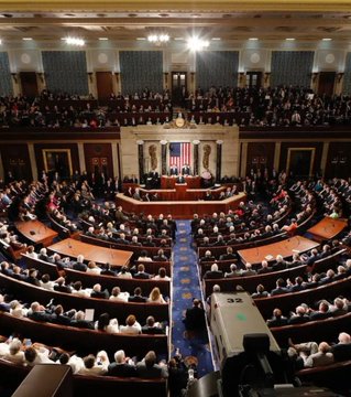 Interior view of the US House of Representatives chamber filled with people, showing the speaker's rostrum with an American flag and seating arranged in a semicircle.