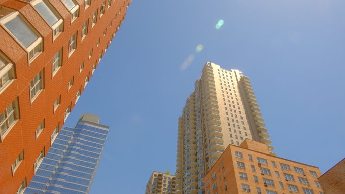 An upward view of tall urban buildings including a red brick high-rise and a beige tower against a clear blue sky with some lens flare.