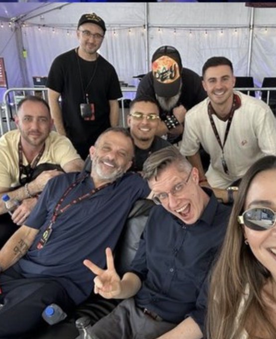 A group photo of seven people posing together, with one man in the front making a peace sign and another wearing distinctive gold/yellow rimmed glasses.