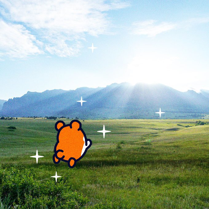 A scenic landscape of a green grassy field with mountains in the background under a bright blue sky, featuring a cartoon illustration of a small orange bear viewed from behind with sparkle effects added.