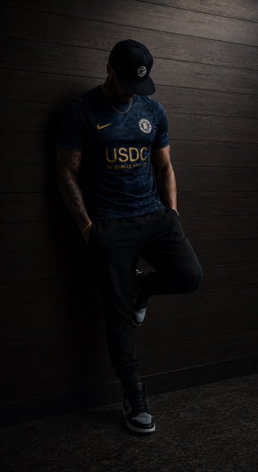 A young man poses in a moody, low-lit photo wearing a navy Chelsea FC jersey featuring the "USDC" sponsor logo and Nike swoosh, paired with a black cap and dark jeans. The image serves as a stylized promotional visual for the registration of the new USDC-sponsored Chelsea jersey mentioned in the post.