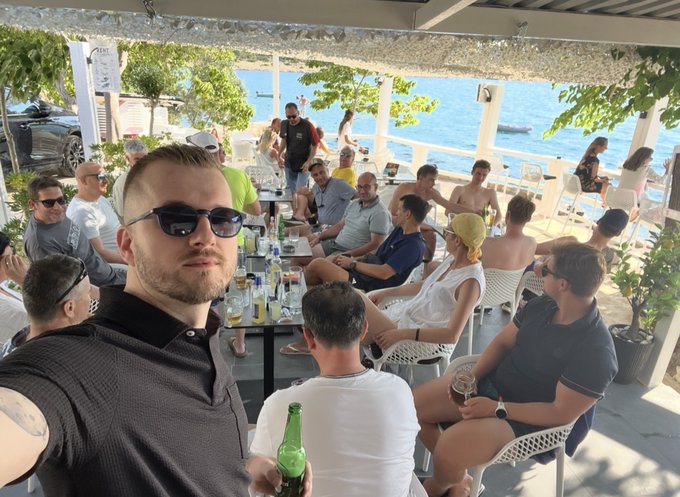 A man in sunglasses takes a selfie at a lively beachside gathering, with a group of people sitting at tables enjoying drinks in the background, countering the "bots" narrative behind the $SONG project by showing real community members who traveled to meet in person.