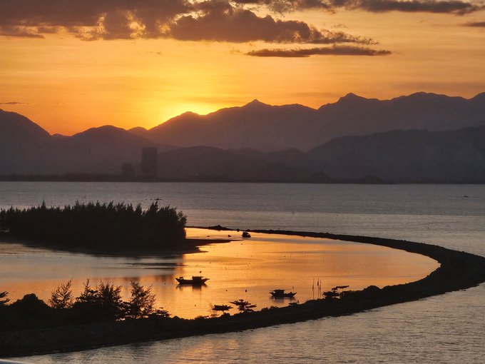 A scenic landscape photograph capturing a vibrant orange sunset over silhouetted mountains, with small boats resting in the calm waters of a curved shoreline.