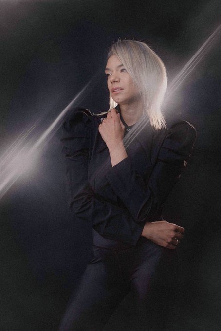 A studio portrait of a blonde woman in a dark outfit posing against a black background with dramatic streaks of light crossing the frame.