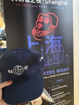 A hand holds up a navy blue "BAYC" (Bored Ape Yacht Club) baseball cap in front of a promotional banner for the "MAKERS NIGHT" co-creators event in Shanghai, with the date "12/20" visible, connecting the swag to the recent Yuga Labs Asia Tour event.