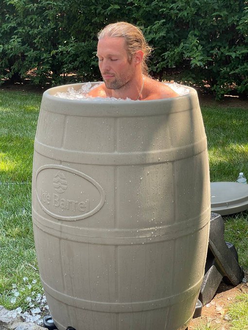 A man with long blonde hair relaxes inside a tan 'Ice Barrel' filled with ice water, set up on a grassy lawn with bushes in the background.