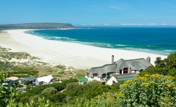 An aerial coastal view showing a long white sandy beach and turquoise ocean waves, with a house featuring multiple gabled roofs in the foreground surrounded by green vegetation.