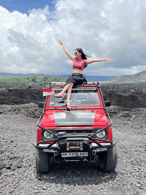 A woman triumphantly raises her arms while sitting atop a red 4x4 Jeep (license plate "DK 1399 AAT") on a rocky volcanic landscape with the Indonesian flag, celebrating her first sponsored event by @thebitmela in the web3 space. The adventurous pose and rugged setting visually convey her excitement and milestone of achieving a sponsored crypto event after years of behind-the-scenes work.