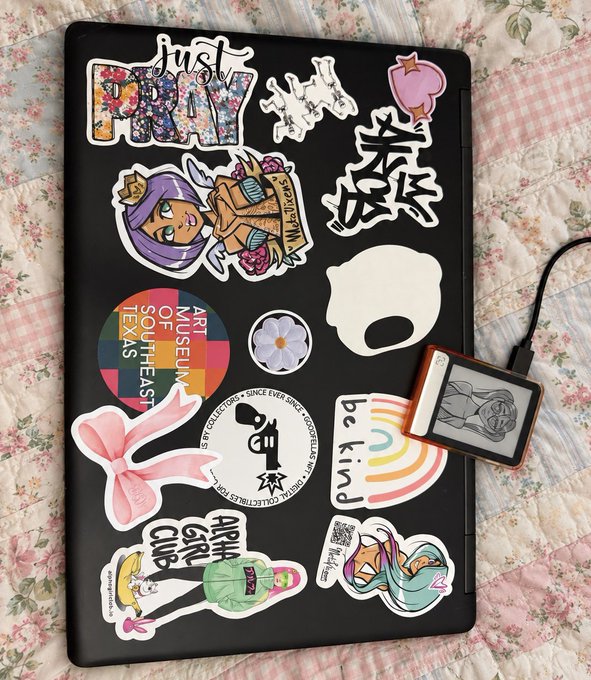 A black laptop covered in various decorative stickers rests on a floral quilted blanket, accompanied by an e-reader device with a cable plugged in.