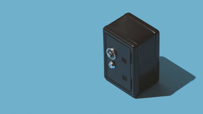 A 3D render shows a small black safe with a combination dial and keyhole sitting on a light blue surface, casting a dark shadow.