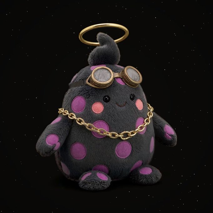 A 3D-rendered, dark grey, egg-shaped plush toy with pink spots, a gold halo, brass goggles, and a gold chain necklace stands against a starry black background.