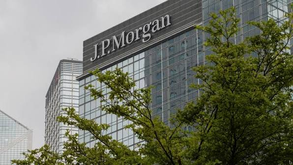 The exterior of a modern glass office building displaying the 'J.P. Morgan' logo, partially obscured by green tree branches in the foreground.