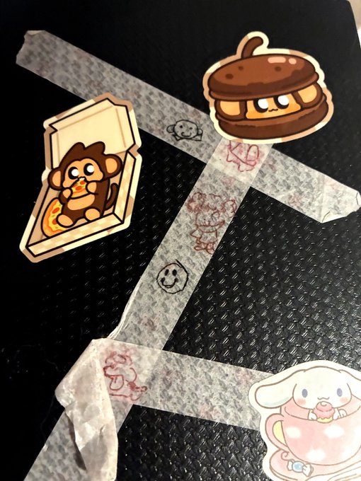Cute cartoon stickers including a chocolate sandwich cookie, a monkey in a cereal box, and a bunny in a teacup decorate a black notebook covered with masking tape and small doodles.