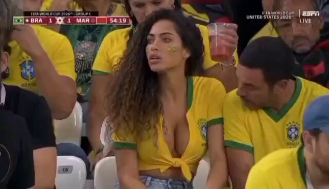 The image shows a woman wearing yellow and green clothing, sitting among other people who appear to be watching a soccer game. She has her hair styled in an updo and is holding a drink. The surrounding crowd seems engaged in the game as well.