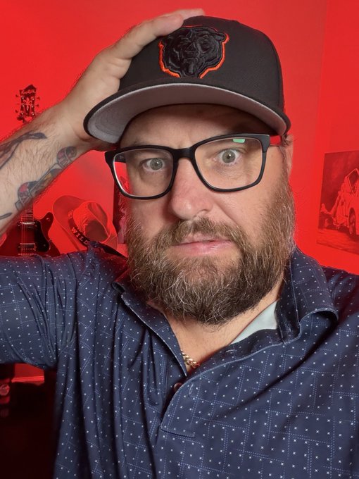 A close-up selfie of a bearded man wearing black glasses and a black cap with a bear logo, adjusting his hat while seated in a room with red lighting and guitars.