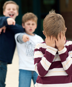 A childhood photo of a young boy in a striped maroon and white shirt covering his face with his hands, while two other boys stand behind him with angry expressions and pointed fingers.