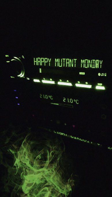 A car dashboard display illuminated in green showing 'HAPPY MUTANT MONDAY' with temperature readings of 21.0°C, with green smoke visible in the foreground.