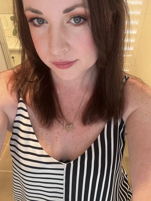 A selfie of a smiling woman with auburn hair and freckles, wearing a black and white striped sleeveless top and a round pendant necklace.