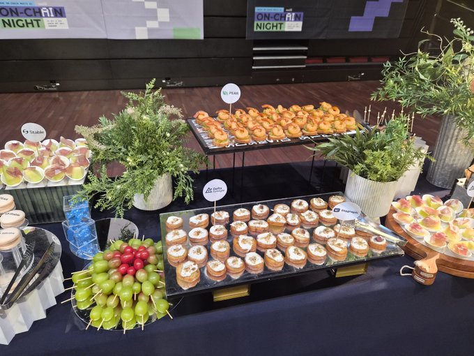A catering spread on a blue table featuring various appetizers including bruschetta, fruit skewers, and canapés, with small labels for 'St-Jean', 'St-Féar', 'Data Decoder', and 'Magnetics', under 'ON-CHAIN NIGHT' banners.