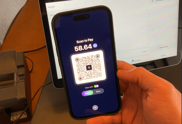 A person holds a smartphone displaying a "Scan to Pay" screen for **$58.64** in USDC, with a QR code offering payment via Coinbase or Base—demonstrating a working prototype of an app that enables in-person USDC transactions for shops. In the background, a point-of-sale terminal screen reinforces the real-world retail use case.